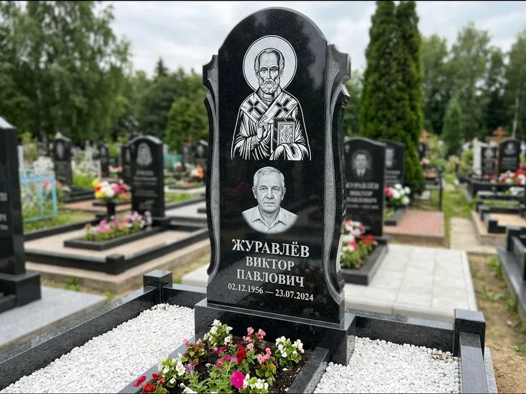 Памятник с гравировкой иконы: стела с образом в полный рост на обороте — гравировка образа Матроны Московской, низкие кусты спиреи в цвету, работа «Лебедева» в Набережных Челнах